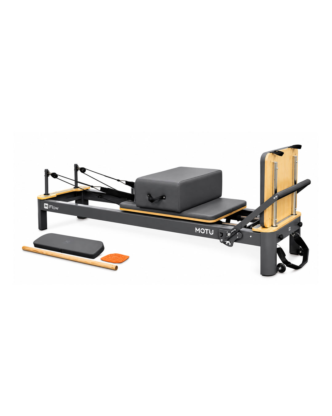A grey MOTU Flow Pilates reformer with maple wood trim, a padded carriage with a grey box on top, rope and pulley risers at one end and an upright footboard at the other. A grey pad, a wooden pole and an orange pad lie in front.