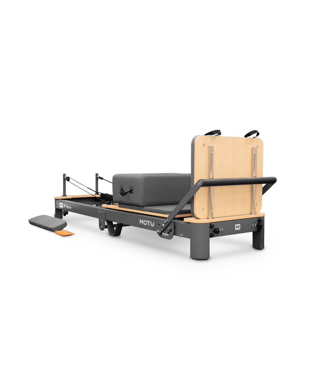 A dark grey folding Pilates reformer with a pale wooden jump board standing upright at one end, a grey padded sitting box on the carriage and a grey foot platform on the floor beside the frame.