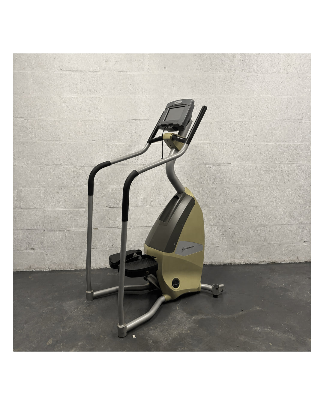 A Stairmaster SC5 stepper with a pale gold and grey body, silver handrails with black grips, black foot pedals and a screen console, standing against a white painted brick wall on a dark grey floor.